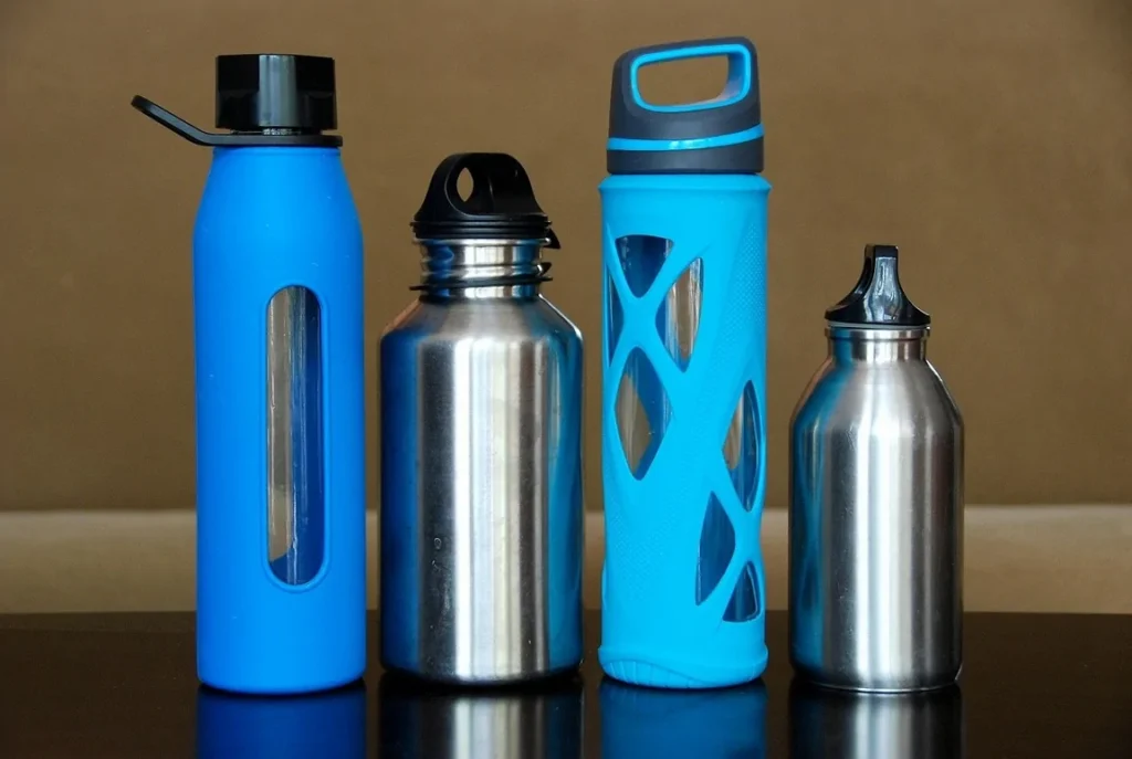 Various types of reusable water bottles made of stainless steel and glass with protective sleeves, standing side by side